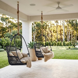 May include: Two black wicker hanging chairs with beige cushions and blankets are suspended from a porch ceiling. The chairs are hanging from thick, knotted ropes. A large potted plant sits between the chairs. The background features a green lawn and trees.