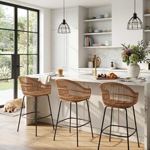 May include: Three bar stools with rounded, woven rattan backs and seats, supported by four slender black metal legs with a circular footrest. The stools are positioned in front of a white marble kitchen island with a gold faucet and decor.