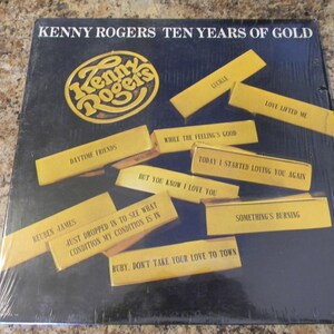 May include: A black vinyl record album cover with a gold background. The album is titled "Kenny Rogers Ten Years of Gold". The cover features a gold circle with the text "Kenny Rogers" inside. The album cover also features a collage of gold rectangles with song titles printed on them, including "Reuben James", "Daytime Friends", "Love Lifted Me", "While the Feeling's Good", "Today I Started Loving You Again", "But You Know I Love You", "Just Dropped In to See What Condition My Condition Is In", "Ruby, Don't Take Your Love to Town", "Something's Burning", and "Lucille".