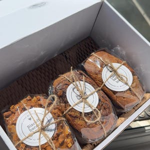 May include: Three small loaves of banana bread packaged in a white cardboard box. Each loaf is wrapped in clear plastic, tied with twine, and features a round white label. The labels read Biscoff banana, Nutella banana, and Classic banana bread.