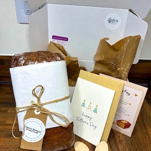 May include: A loaf of banana bread wrapped in plastic and white paper with twine and a tag reading Classic Banana Bread Loaf, accompanied by a Father's Day card, a note from Loaf by Fay, and two wooden spoons inside an open white cardboard box.