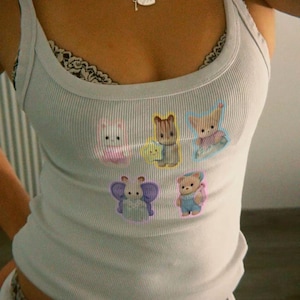 May include: A light blue ribbed tank top with cartoon animal graphics. The tank top has a scoop neckline and thin straps. A silver necklace with a heart-shaped pendant is visible. The top is paired with white bottoms.