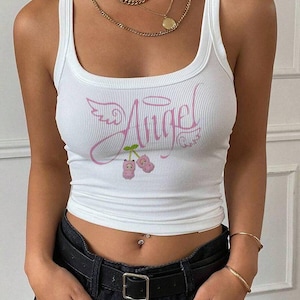 Op de afbeelding: Een wit geribbelde tanktop met een roze grafische print op de borst. Het ontwerp bevat het woord Angel in cursief schrift, vergezeld van een halo, vleugels en twee kleine roze kersachtige figuren die aan een groen steeltje hangen.