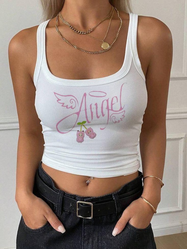 Op de afbeelding: Een wit geribbelde tanktop met een roze grafische print op de borst. Het ontwerp bevat het woord Angel in cursief schrift, vergezeld van een halo, vleugels en twee kleine roze kersachtige figuren die aan een groen steeltje hangen.