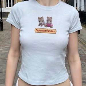 May include: A light blue short-sleeved t-shirt featuring a graphic of two small cat figures and the text Sylvanian Families printed in a red-bordered yellow rectangular box centred on the chest area of the garment.