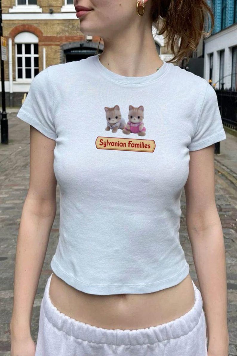 May include: A light blue short-sleeved t-shirt featuring a graphic of two small cat figures and the text Sylvanian Families printed in a red-bordered yellow rectangular box centred on the chest area of the garment.