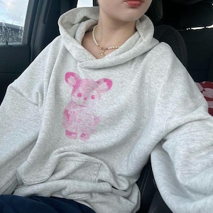 May include: A light gray heathered hoodie featuring a long-sleeved design with a hood and a front kangaroo pocket. The center of the chest displays a pink graphic print of a stylized bear figure with rounded ears and a textured appearance.