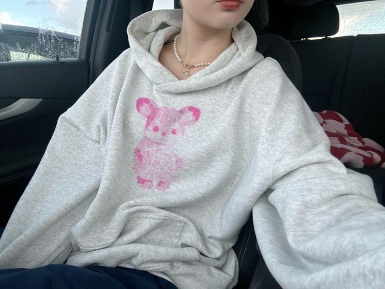 May include: A light gray heathered hoodie featuring a long-sleeved design with a hood and a front kangaroo pocket. The center of the chest displays a pink graphic print of a stylized bear figure with rounded ears and a textured appearance.