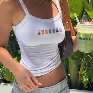 May include: White tank top with thin straps and a row of colorful cartoon characters across the chest. The top is paired with gray sweatpants. A small brown purse with a gold chain strap is visible.