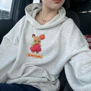 May include: A light gray hoodie with a kangaroo pocket and a drawstring hood. The hoodie features a graphic of a Sylvanian Families character holding a tomato. The text "Sylvanian Families" is below the graphic.