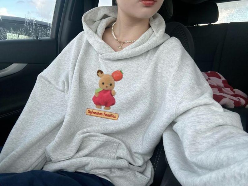 May include: A light gray hoodie with a kangaroo pocket and a drawstring hood. The hoodie features a graphic of a Sylvanian Families character holding a tomato. The text "Sylvanian Families" is below the graphic.