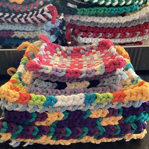 May include: Stacks of colorful, handmade pot holders in various patterns and colors. The pot holders are made of woven yarn in shades of green, red, white, purple, yellow, and blue. Each pot holder has a loop for hanging.