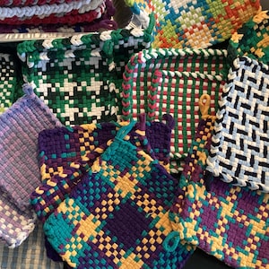 May include: A collection of colorful, woven pot holders in various patterns and colors, including green, purple, yellow, and blue. Each pot holder has a loop for hanging. The pot holders are handmade and appear to be made of yarn or fabric.