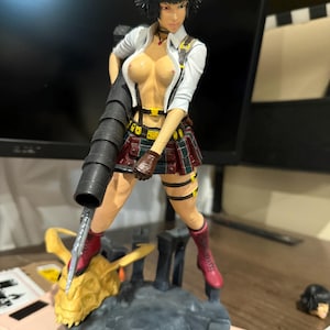 Puede incluir: Figura detallada de un personaje femenino de videojuego, con camisa blanca, falda a cuadros y botas rojas. Sostiene un arma grande y está de pie sobre una base detallada con una cabeza de dragón dorada. La figura tiene cabello negro y está en una pose dinámica.