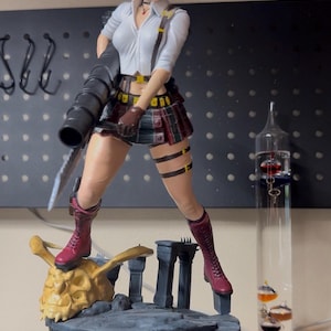 Puede incluir: Figura detallada de un personaje femenino de videojuego, de pie sobre una base de piedra. Viste camisa blanca, falda a cuadros y botas rojas, sosteniendo un arma grande. La figura es muy detallada y tiene una pose dinámica.