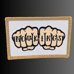 May include: A rectangular embroidered patch with a white background and a tan border. The design features two tan fists with black outlines, each displaying letters across the knuckles that spell out the text NO KINGS with a crown icon.