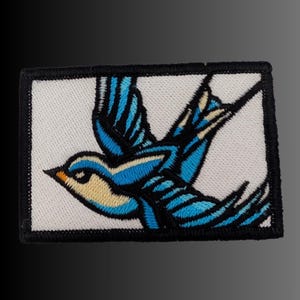 May include: A rectangular embroidered patch featuring a stylized bird in flight. The bird has bright blue wings with black outlines, a cream-colored chest, and a small orange beak. The entire design is set against a white background with a thick black border.