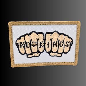 May include: A rectangular embroidered patch with a white background and a tan border. The design features two tan fists with black outlines, each displaying letters across the knuckles that spell out the text NO KINGS with a crown icon.