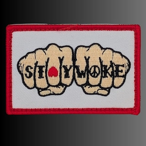 May include: A rectangular embroidered patch with a white background and a red border. The design features two beige fists side by side with the black text STAY WOKE across the knuckles, incorporating a red heart and a black peace symbol.