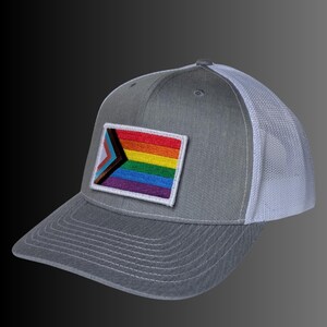 May include: A gray baseball cap with a white mesh back and a rectangular patch on the front. The patch features a horizontal rainbow pride flag design with a black and brown chevron triangle on the left side, stitched with white borders.