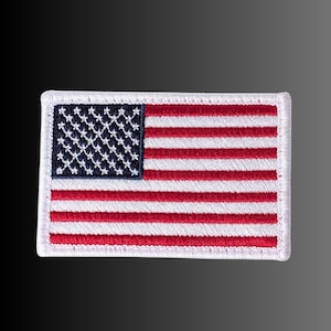May include: A rectangular embroidered patch featuring the United States flag. The design includes a dark blue canton with white stars and alternating horizontal red and white stripes, all bordered by a thick white stitched edge finish.