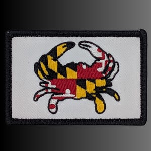 May include: A rectangular embroidered patch featuring a white background and a black border. The center displays a crab graphic filled with the Maryland state flag pattern, consisting of alternating yellow, black, red, and white geometric sections.