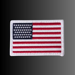 May include: A rectangular embroidered patch featuring the United States flag. The design includes a dark blue canton with white stars in the upper left corner, alongside horizontal red and white stripes. The entire patch has a white stitched border.
