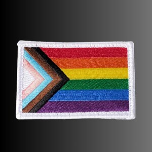May include: A rectangular embroidered patch featuring the Progress Pride flag design. The patch displays horizontal stripes of red, orange, yellow, green, blue, and violet, with a chevron on the left side in white, pink, light blue, brown, and black.