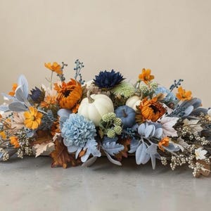 May include: A floral arrangement featuring white and blue pumpkins nestled among orange, blue, and white artificial flowers, dusty miller leaves, brown autumn foliage, and small white berries, creating a dense, horizontal decorative centerpiece.