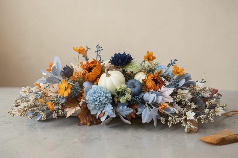 May include: A floral arrangement featuring white and blue pumpkins nestled among orange, blue, and white artificial flowers, dusty miller leaves, brown autumn foliage, and small white berries, creating a dense, horizontal decorative centerpiece.