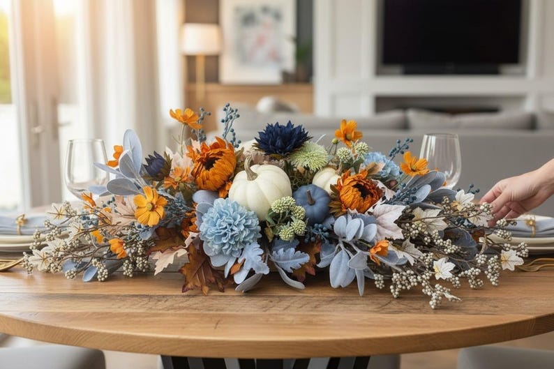 May include: A floral centerpiece featuring white and blue pumpkins, orange sunflowers, light blue chrysanthemums, dark blue dahlias, dusty miller foliage, and white berry sprigs arranged on a wooden table with two clear wine glasses.