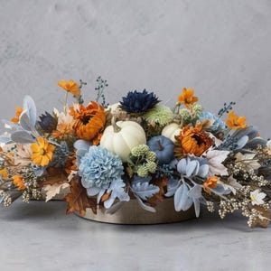May include: A floral centerpiece featuring white and blue pumpkins nestled among orange, blue, and white flowers, dusty miller leaves, and small berries. The arrangement is set in a low, circular container, creating a wide, horizontal display.