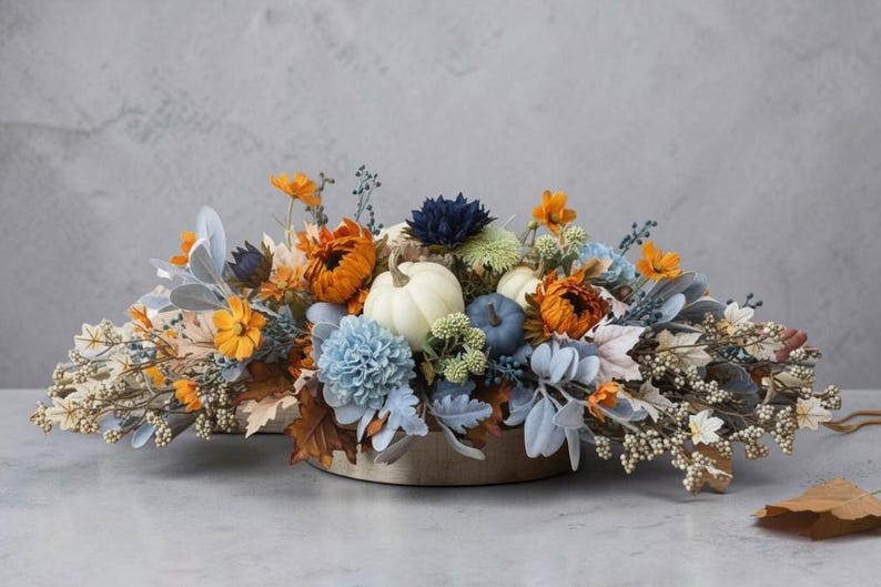 May include: A floral centerpiece featuring white and blue pumpkins nestled among orange, blue, and white flowers, dusty miller leaves, and small berries. The arrangement is set in a low, circular container, creating a wide, horizontal display.