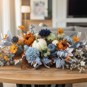May include: A floral centerpiece featuring white and blue pumpkins, orange sunflowers, light blue chrysanthemums, dark blue dahlias, dusty miller foliage, and white berry sprigs arranged on a wooden table with two clear wine glasses.