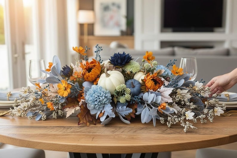May include: A floral centerpiece featuring white and blue pumpkins, orange sunflowers, light blue chrysanthemums, dark blue dahlias, dusty miller foliage, and white berry sprigs arranged on a wooden table with two clear wine glasses.