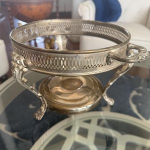 May include: Silver-toned metal serving dish with a mirrored interior. The dish has a decorative pierced rim and ornate handles. It is supported by three curved legs and a central base, showcasing a vintage aesthetic.