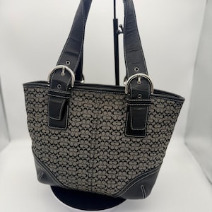 May include: A black and gray tote bag with a repeating floral pattern. The bag has black leather handles and trim, with silver-colored buckles. White stitching accents the leather details. The bag is sitting on a white surface.