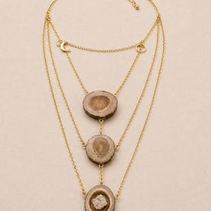 May include: A layered gold-tone necklace featuring three strands. Each strand has a different pendant: a crescent moon, a small ring, and three circular pendants made of a light brown material with a natural, textured appearance. The necklace is displayed on a neutral background.
