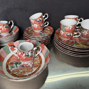 May include: A collection of vintage porcelain teacups, saucers, and serving dishes. The set features a vibrant design with red, green, and gold accents, including a bird and floral motif. The teacups have gold handles, and the saucers are approximately 6 inches in diameter.