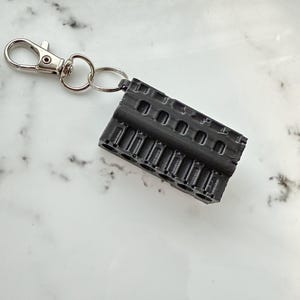 May include: A black, 3D-printed keychain featuring a detailed engine block design. The keychain includes a silver-colored clasp and a split ring for attaching keys. The engine block is rectangular with multiple cylinders and intricate detailing.