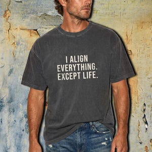 May include: A charcoal grey short-sleeve t-shirt featuring the white text I ALIGN EVERYTHING. EXCEPT LIFE. printed in a distressed font on the chest. The garment has a crew neckline and a relaxed fit, paired with blue denim jeans.