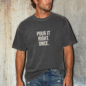 May include: A charcoal gray short-sleeve t-shirt with a crew neckline. The front of the shirt features the text "POUR IT RIGHT. ONCE." printed in a distressed, off-white sans-serif font centered on the chest area of the garment.