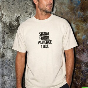 May include: A cream-colored short-sleeve t-shirt featuring the black text "SIGNAL FOUND. PATIENCE LOST." printed in a distressed, sans-serif font on the center of the chest. The garment has a crew neckline and a standard, relaxed fit silhouette.