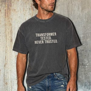 May include: A charcoal gray short-sleeve t-shirt with a crew neckline. The front of the shirt features the white text "TRANSFORMER TESTED. NEVER TRUSTED." printed in a distressed, sans-serif font across the center of the chest.