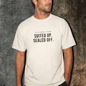 May include: A cream-colored short-sleeve t-shirt featuring the black text "ABATEMENT CREW" at the top and "SUITED UP. SEALED OFF." printed in a bold, distressed sans-serif font across the center of the chest area.