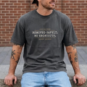 May include: A charcoal gray short-sleeve t-shirt featuring the text "LEAD ABATEMENT REMOVED SAFELY. NO SHORTCUTS. CLEANED. CERTIFIED." printed in white and tan lettering across the chest area in a clean, sans-serif font style.