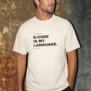 May include: A cream-colored short-sleeve t-shirt featuring black text that reads CNC OPERATOR G-CODE IS MY LANGUAGE. The shirt has a crew neckline and a standard fit, with the graphic printed in a bold, sans-serif font on the chest area.