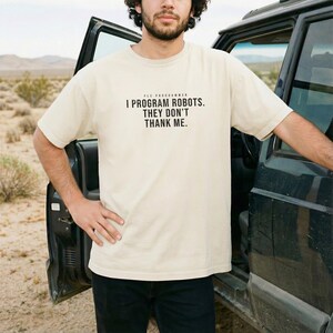 May include: A cream-colored short-sleeve t-shirt featuring black text centered on the chest that reads: PLC PROGRAMMER I PROGRAM ROBOTS. THEY DON'T THANK ME. The shirt has a crew neckline and a standard fit, made of a solid, opaque fabric.