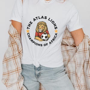 以下が含まれることがあります： サッカーボールを持ったライオンの円形グラフィックが特徴の白いTシャツ。「THE ATLAS LIONS CHAMPIONS OF AFRICA」の文字が黒と金で書かれています。青いジーンズとチェックのシャツと合わせて着用されています。