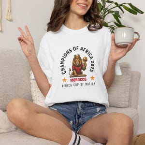 以下が含まれることがあります： 王冠をかぶったライオンがトロフィーを持っているグラフィックの白いTシャツ。「CHAMPIONS OF AFRICA 2025」と「MOROCCO AFRICA CUP OF NATION」の文字。デザインは赤、金、黒。